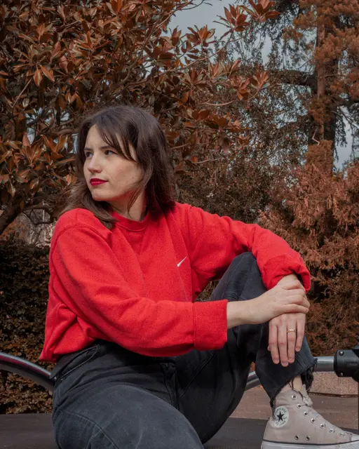 Portrait d'une jeune femme en sweat rouge assise sur un banc rond, devant des arbres aux feuilles rousses.